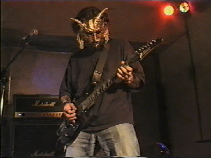 Küçük bir sahnede Marshall amfi ve spot ışıkları önünde gitar çalan boynuzlu maskeli Vesper Aurum